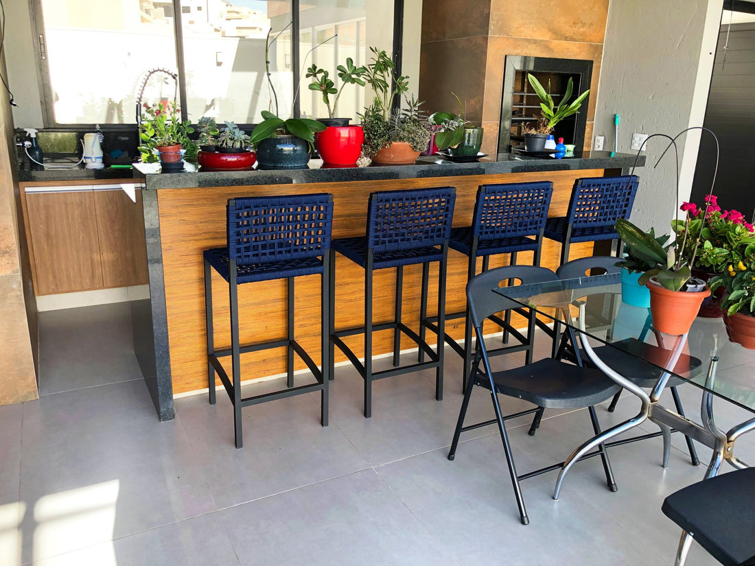 Quatro banquetas de corda náutica azul sobre estrutura de alumínio preto numa bancada de varanda gourmet com churrasqueira, plantas e o lado aberto ao tempo