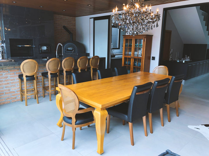 Sala de jantar com mesa grande de madeira maciça, dez cadeiras em volta, banquetas no balcão ao fundo e lustre de cristal acima da mesa
